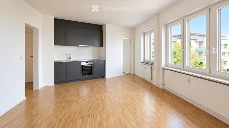 Empty room with wooden flooring, white walls, and large windows. Kitchen area with cabinets and appliances visible in the background.