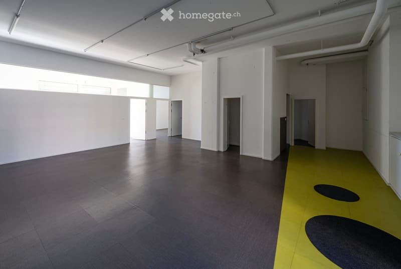 Empty room with grey tiled floor, white walls, large windows, and several doors leading to other spaces. The floor features a yellow section with black circular designs.