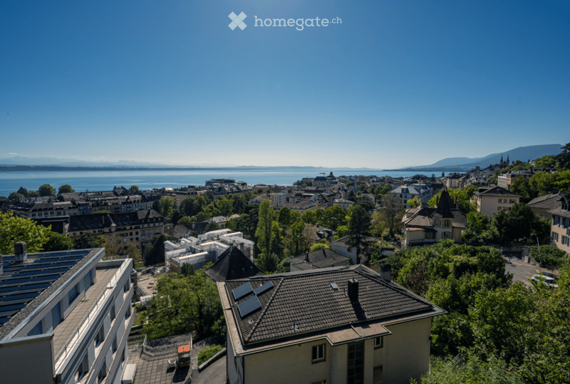 A panoramic view of a cityscape with a lake and mountains in the background, featuring multiple buildings and greenery.