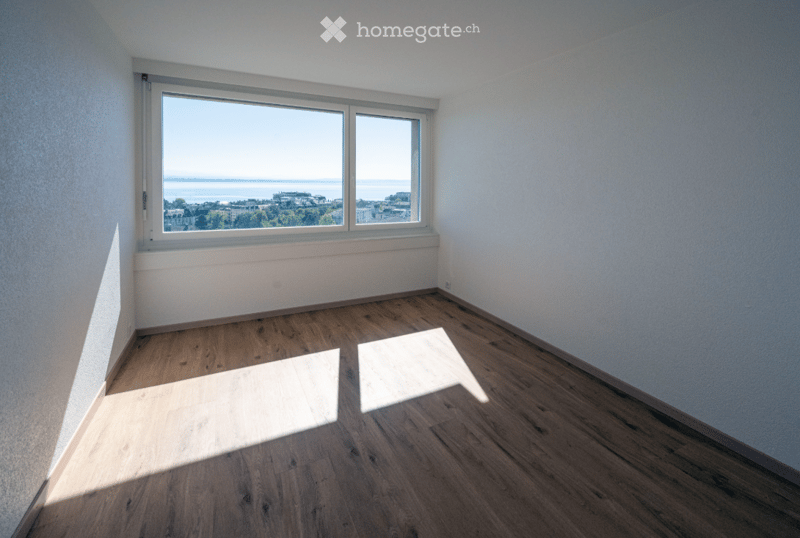 Empty room with wooden flooring and large windows offering a scenic view of the sea and cityscape.