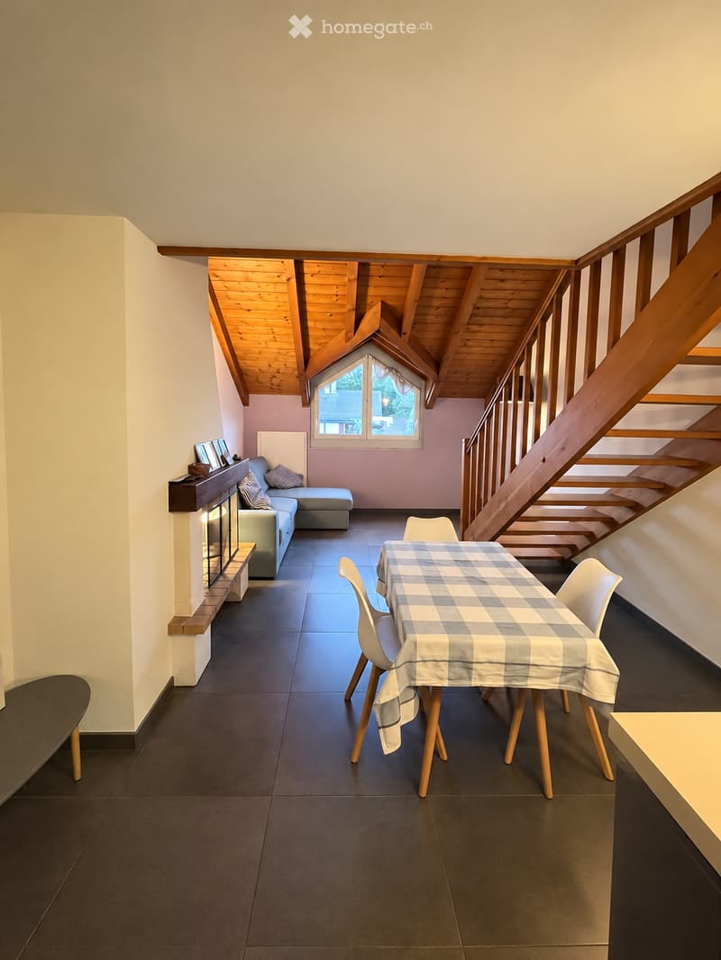 Bright living room with a wooden slanted ceiling, dark tiled floor, and a cozy seating area featuring a sofa, ottoman, and fireplace. A dining table with chairs is set with a checkered tablecloth, and a staircase leads upwards.