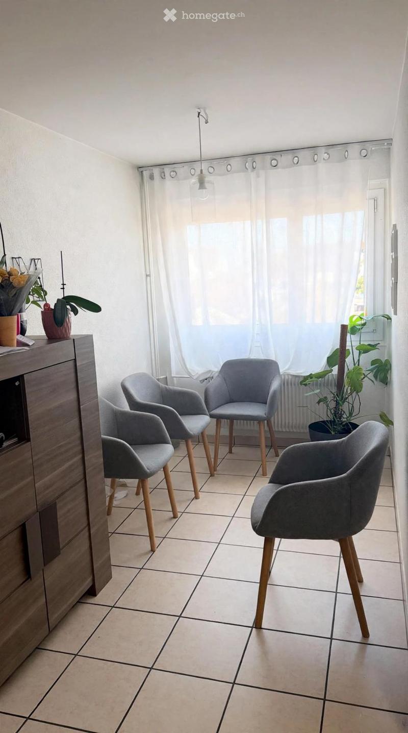 A small, well-lit room with tiled flooring and white walls. It features four modern grey chairs with wooden legs arranged in a semi-circle, a wooden cabinet with a drawer, and several potted plants near the window. The room has large windows covered with