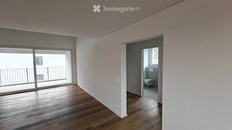Empty room with wooden flooring, white walls, and a sliding glass door leading to a balcony. A small bathroom is visible through an open doorway.