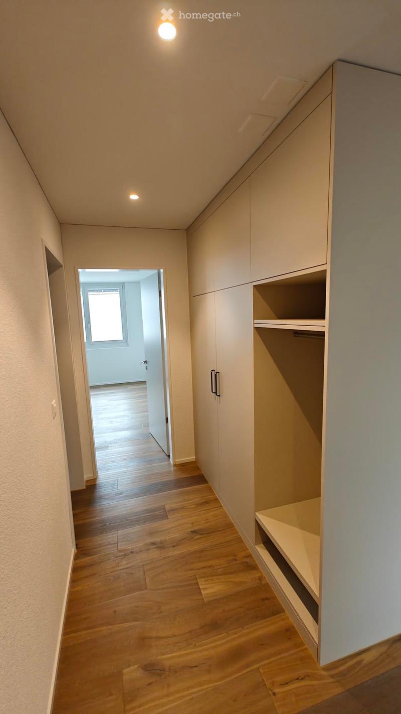 A modern hallway with wooden flooring, built-in white closet with shelves, and recessed lighting. An open doorway leads to another room with a window.
