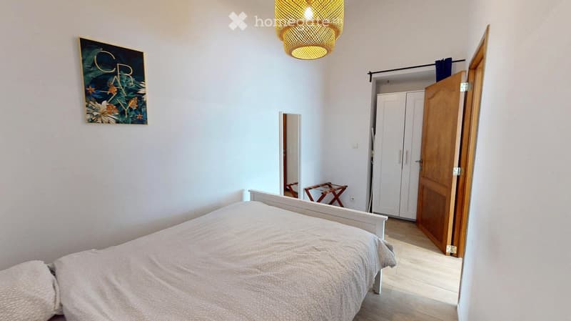 A modern bedroom with a white bed, a wooden wardrobe, a small wooden stool, a decorative picture frame, and a stylish wicker pendant light.