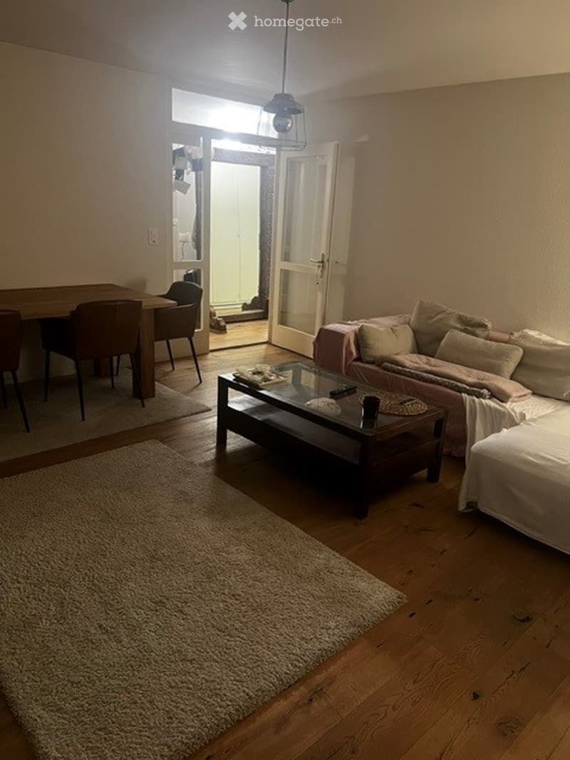 A cozy living and dining area with a sofa, coffee table, dining table, chairs, and a bed, featuring wooden flooring and a large open doorway leading outside.