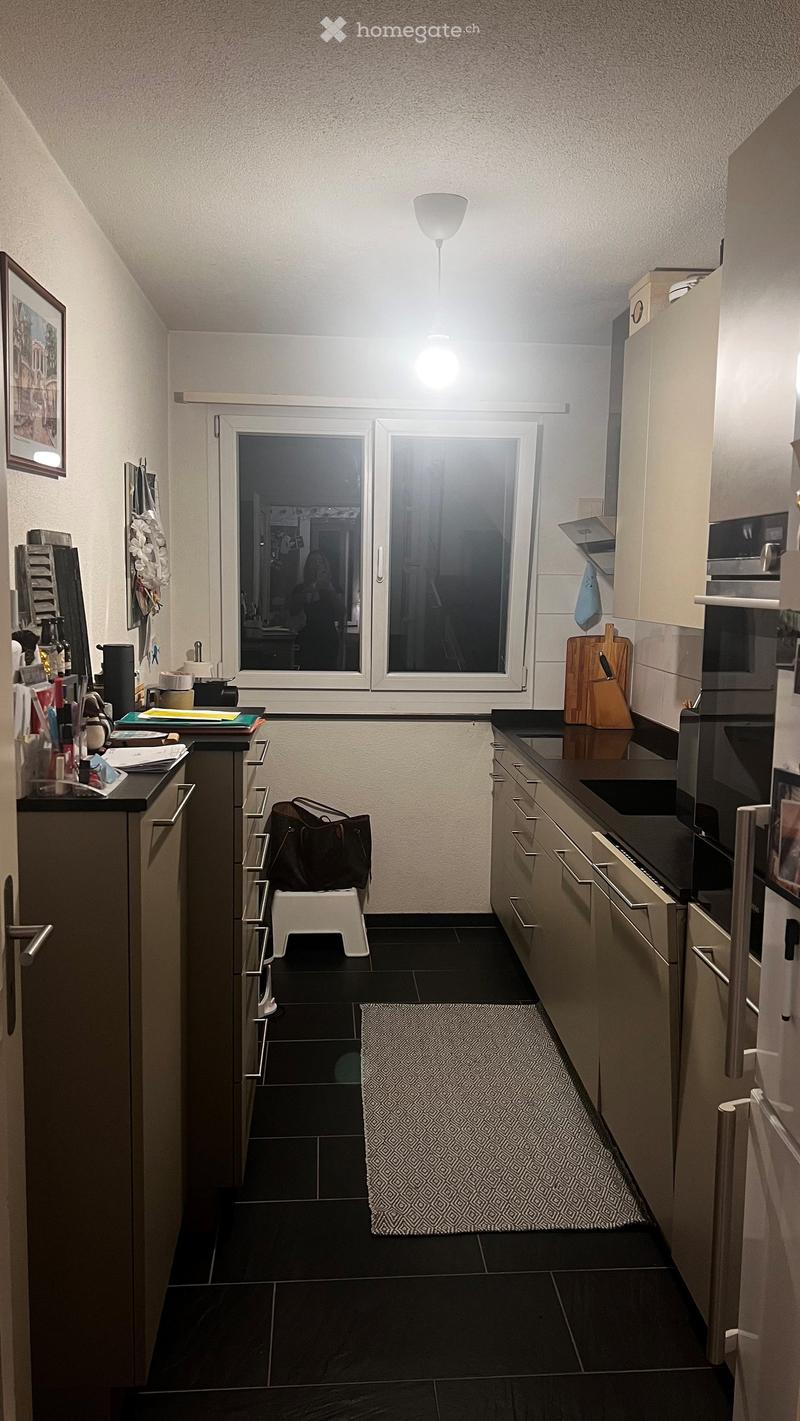 Modern kitchen with light-colored cabinets, black countertops, and a black tiled floor. Features include a refrigerator, oven, microwave, cutting board, stool, handbag, and a framed picture on the wall.