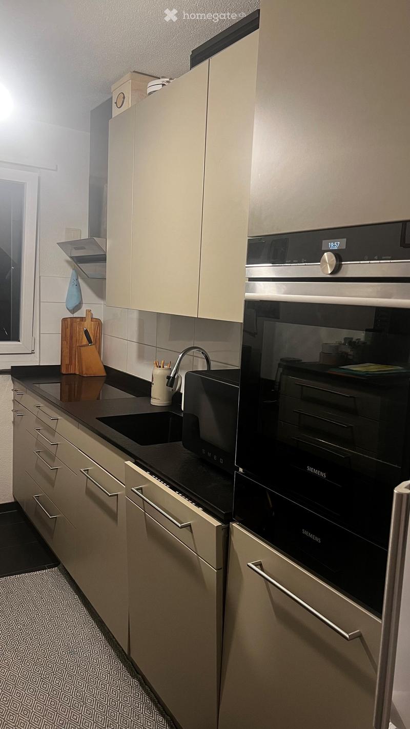 Modern kitchen with light-colored cabinets, black countertops, and stainless steel appliances including an oven and a sink with a modern faucet. A wooden cutting board and a cup are placed on the counter.