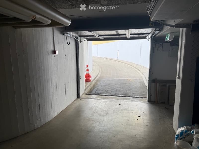 Corridor / Hallway. A narrow, concrete-lined corridor with a curved ceiling and visible pipes. The corridor has a single doorway leading to an open area with traffic cones placed inside.
