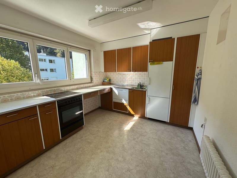 Bright kitchen with wooden cabinets, white refrigerator, oven, sink, and dishwasher. Large windows provide natural light.