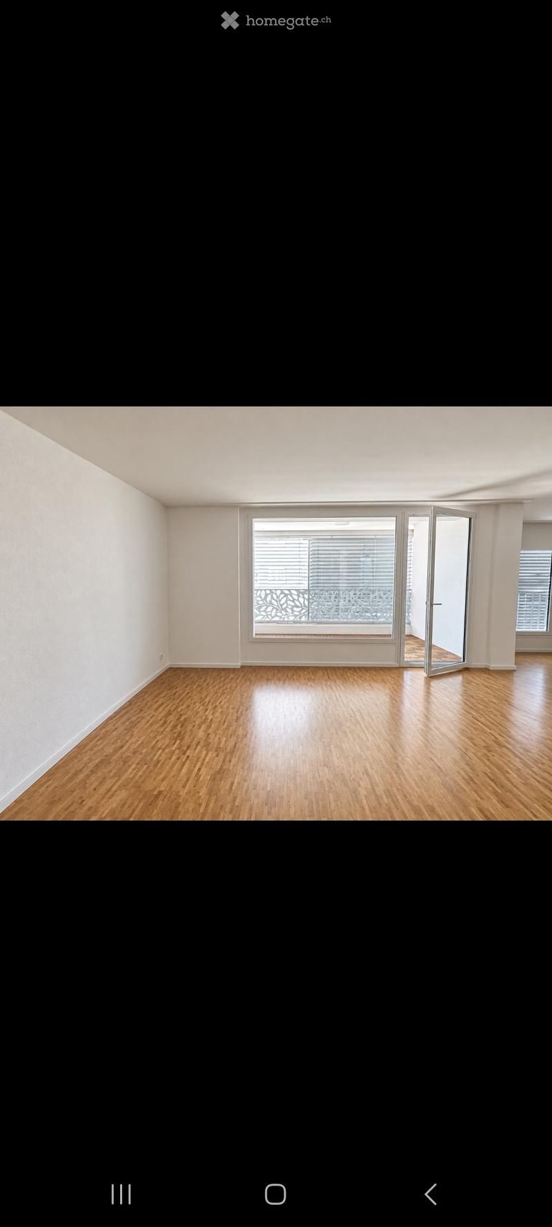 Empty room with wooden flooring, white walls, and large windows with blinds. The room has a sliding glass door leading to another space.