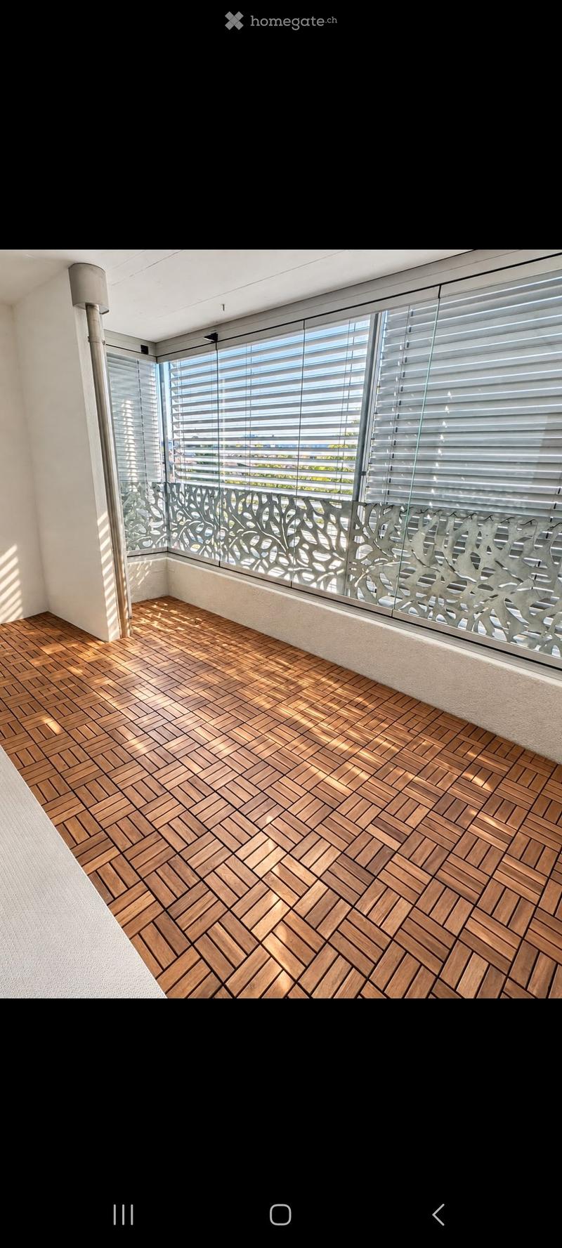 Balcony with wooden herringbone flooring, large windows with blinds, and decorative metal railing.