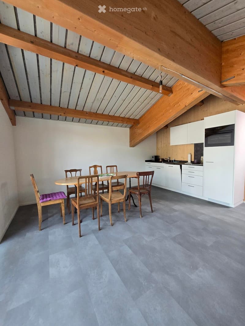Open-plan dining and kitchen area with exposed wooden beams, grey flooring, and modern white cabinets.