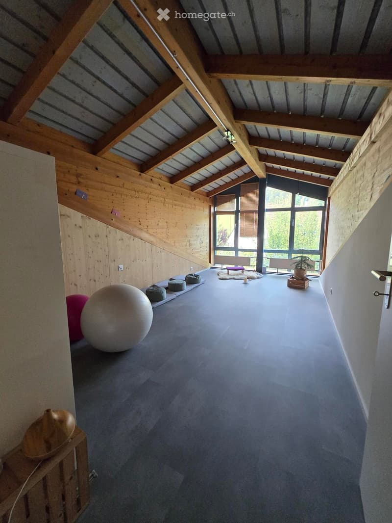 Open, high-ceilinged room with wooden beams, grey carpet, large window with blinds, and scattered exercise equipment including a large exercise ball and small benches.
