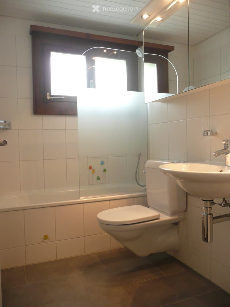 Modern bathroom with white tiles, a toilet, sink, mirror, and bathtub. Features a frosted glass shower enclosure with decorative floral tiles and a window for natural light.
