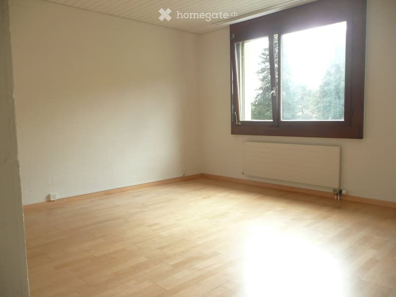 Empty room with wooden flooring, white walls, and a large window with wooden frame. A radiator is mounted on the wall below the window.