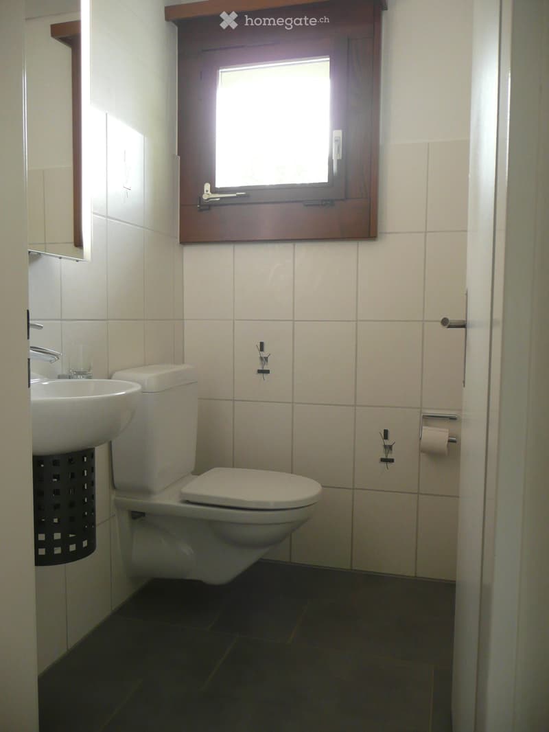A small bathroom with white tiled walls, a toilet, a sink, and a window with wooden frame. The floor is dark tiled.