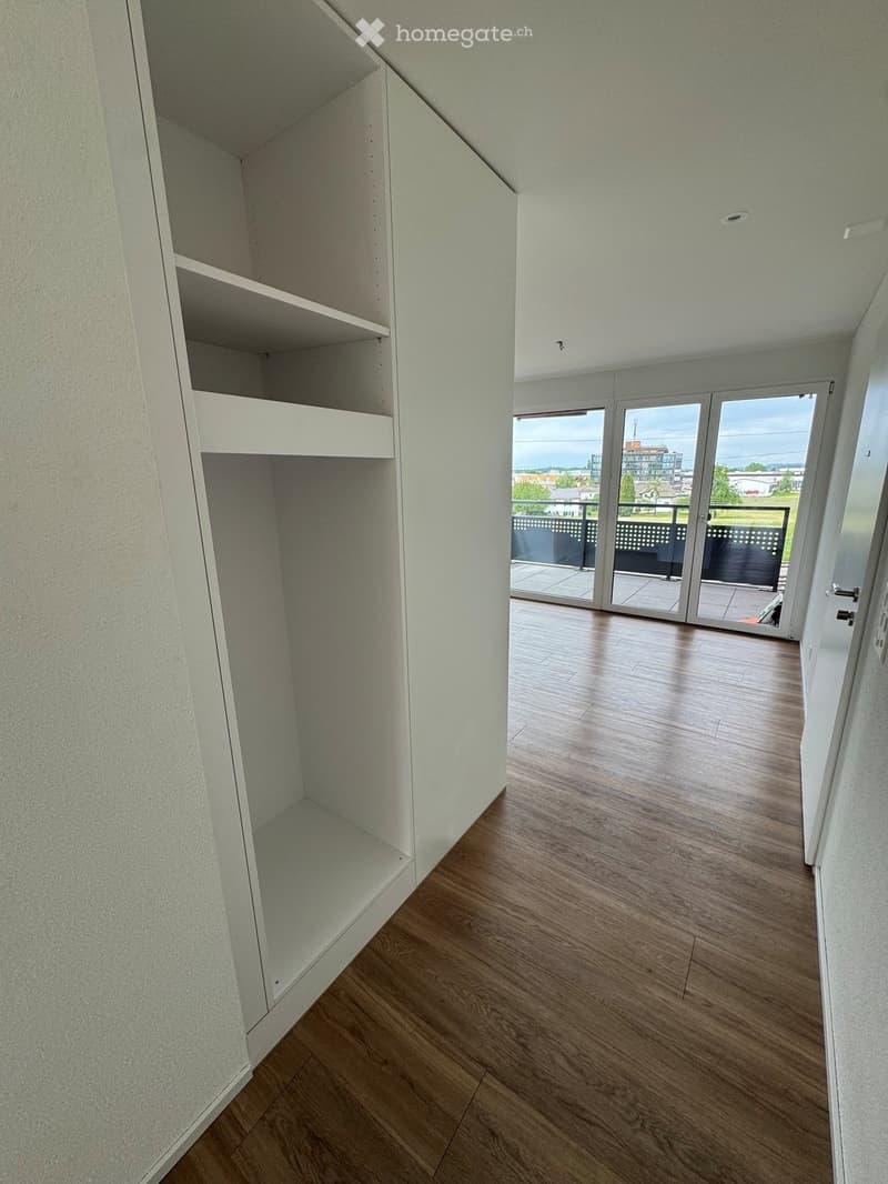 Empty hallway with wooden flooring, white walls, built-in shelving unit, and large sliding glass doors leading to a balcony with a view.