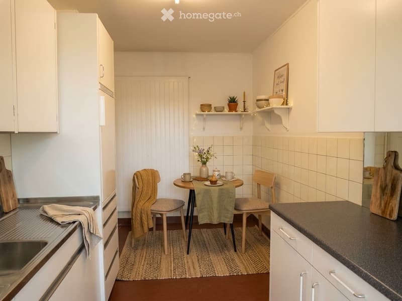 Cozy kitchen with white cabinets, a small dining area with a table and chairs, and a sink. Shelves hold decorative items and plants.