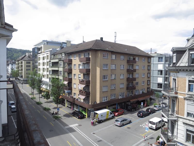 A multi-story residential building with balconies and awnings, located on a street with parked cars and pedestrians.