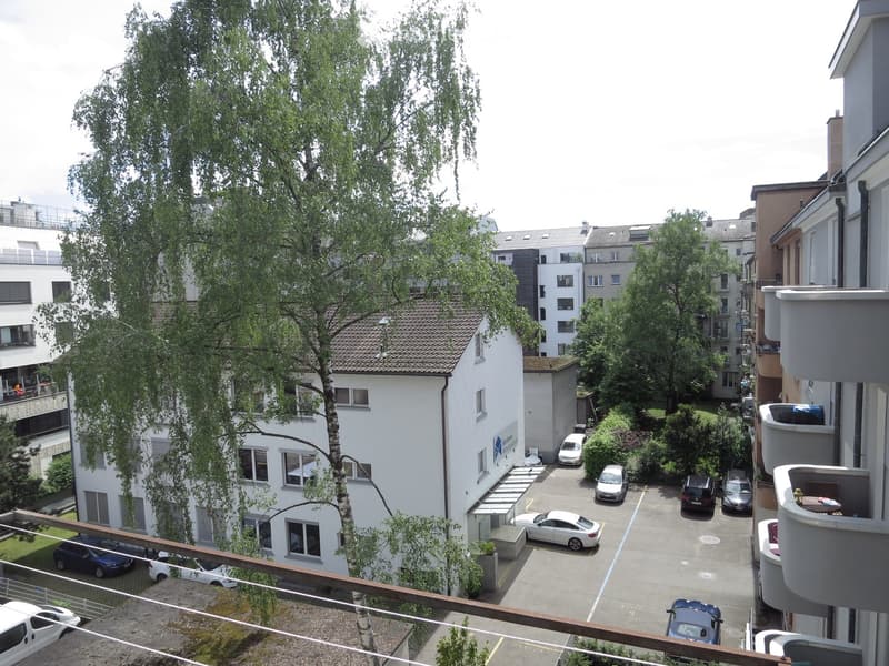 Balcony view overlooking a parking area with several parked cars, surrounded by residential buildings and trees.
