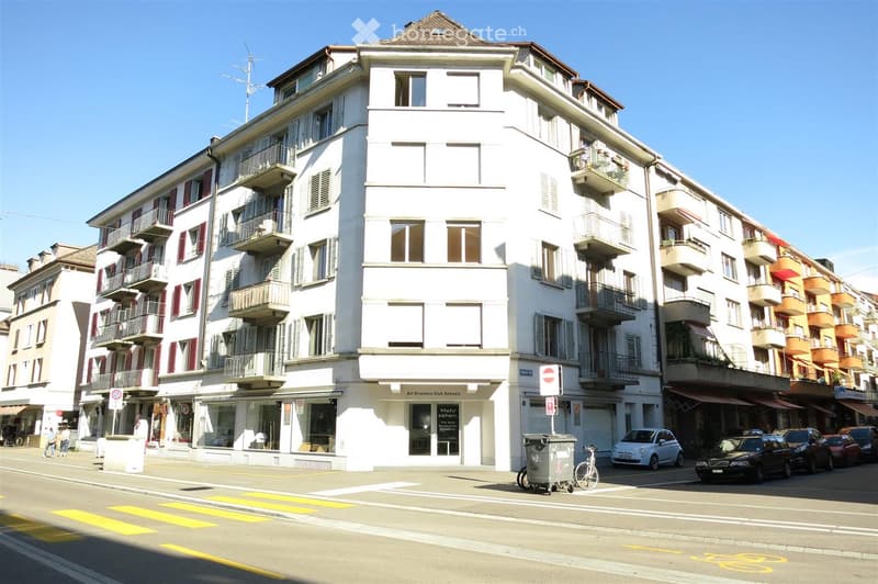 Multi-story white building with balconies and shutters, located on a street with parked cars and pedestrians.