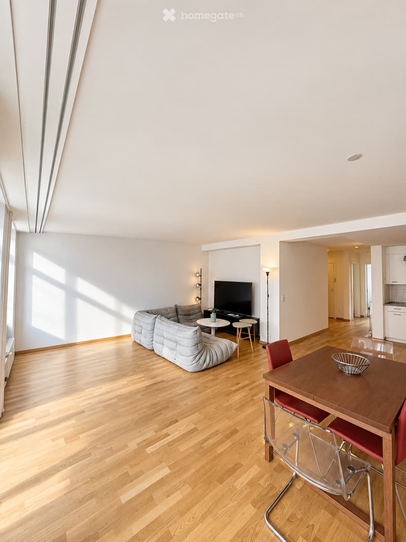 Open-plan living and dining area with light wooden flooring, a grey sectional sofa, a coffee table, a TV, a dining table with chairs, and a floor lamp.