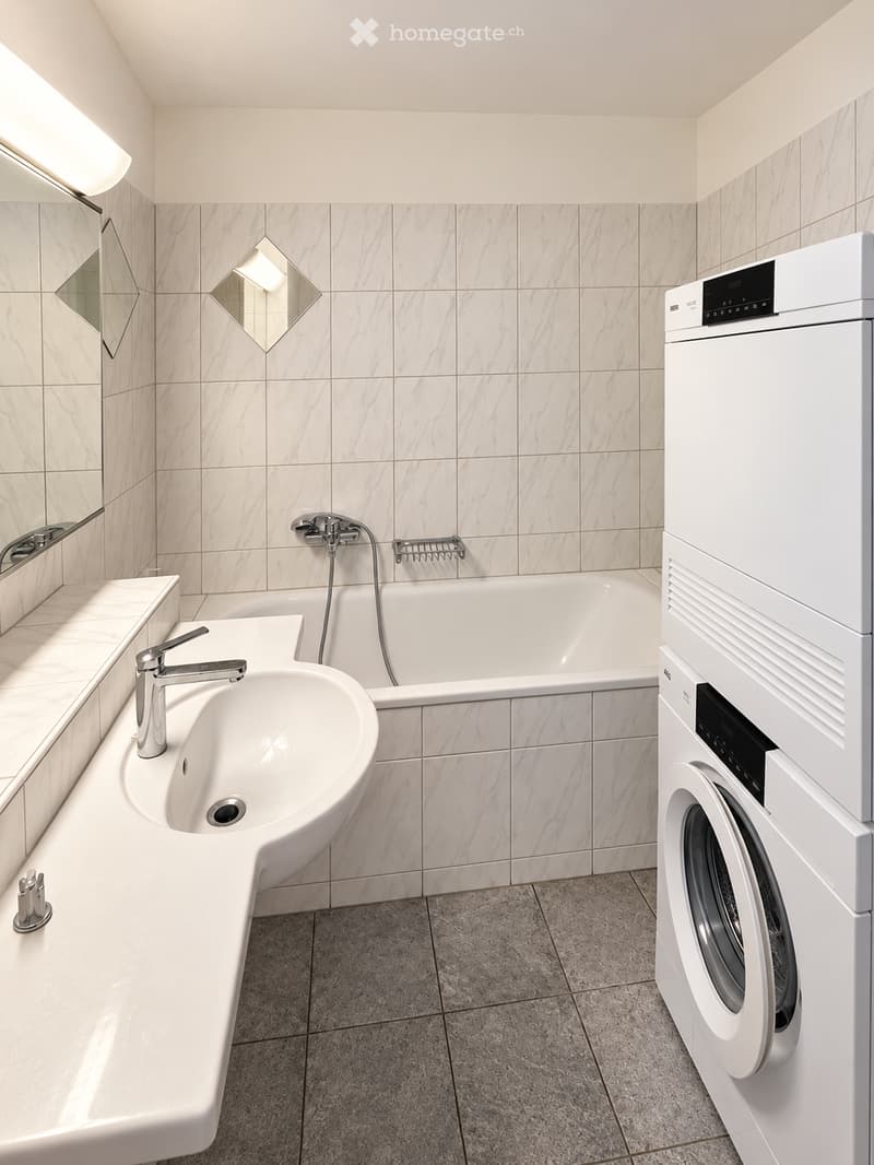 Modern bathroom with marble tiles, a white sink, bathtub, and a stacked washer/dryer unit.