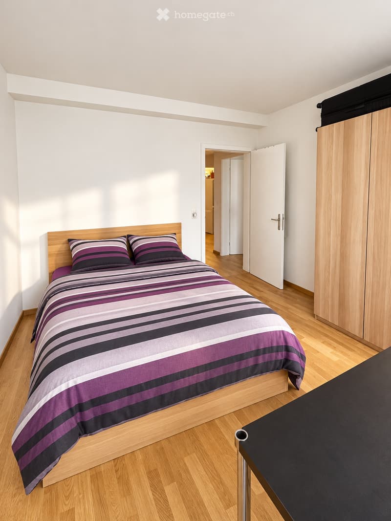 Modern bedroom with wooden flooring, a bed with a purple and grey striped duvet, two matching pillows, and a wooden wardrobe.
