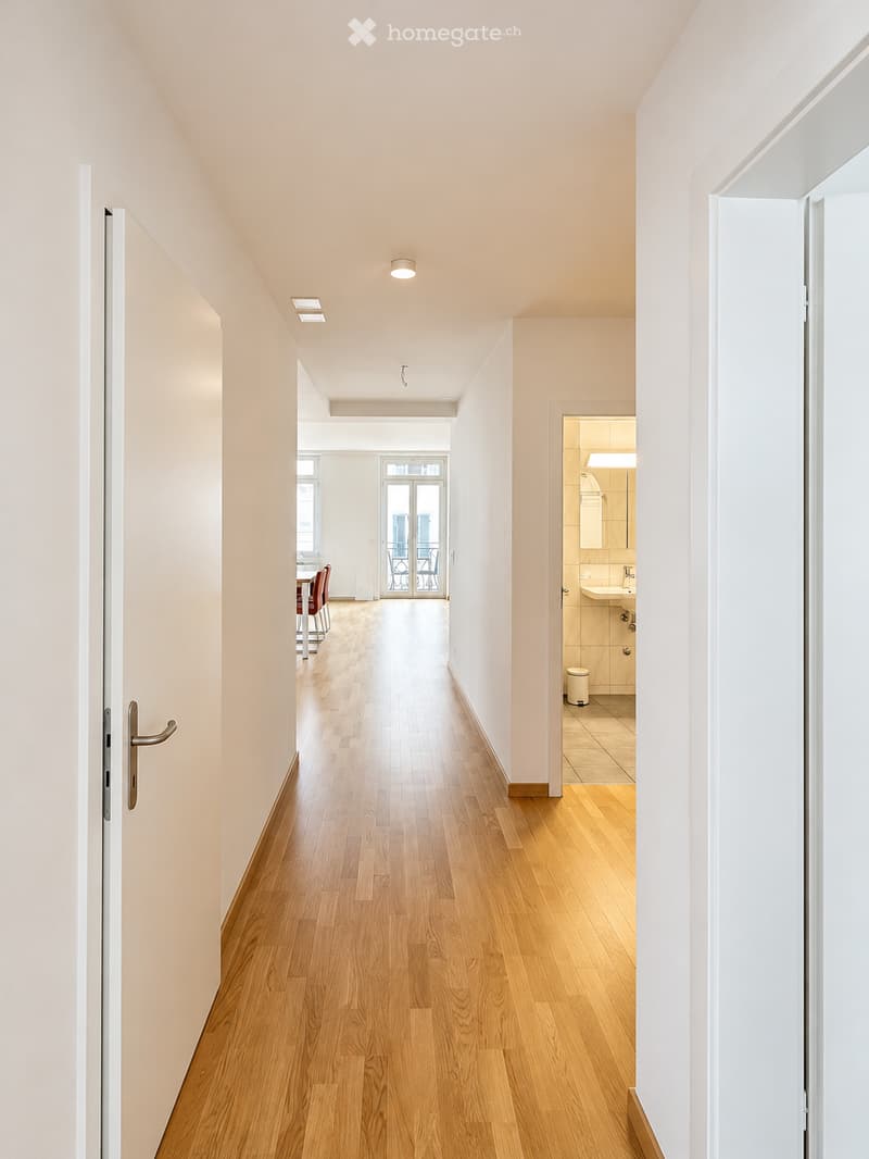 Bright hallway with wooden flooring, white walls, and recessed lighting. Doors lead to a dining area and a bathroom.