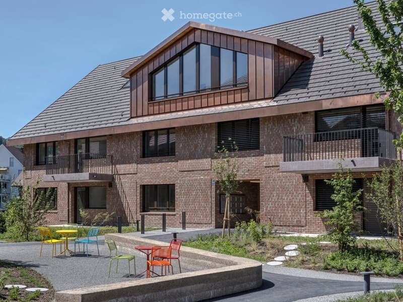 Modern two-story building with brick facade, large windows, and balconies. Outdoor seating area with tables and chairs is visible in front.