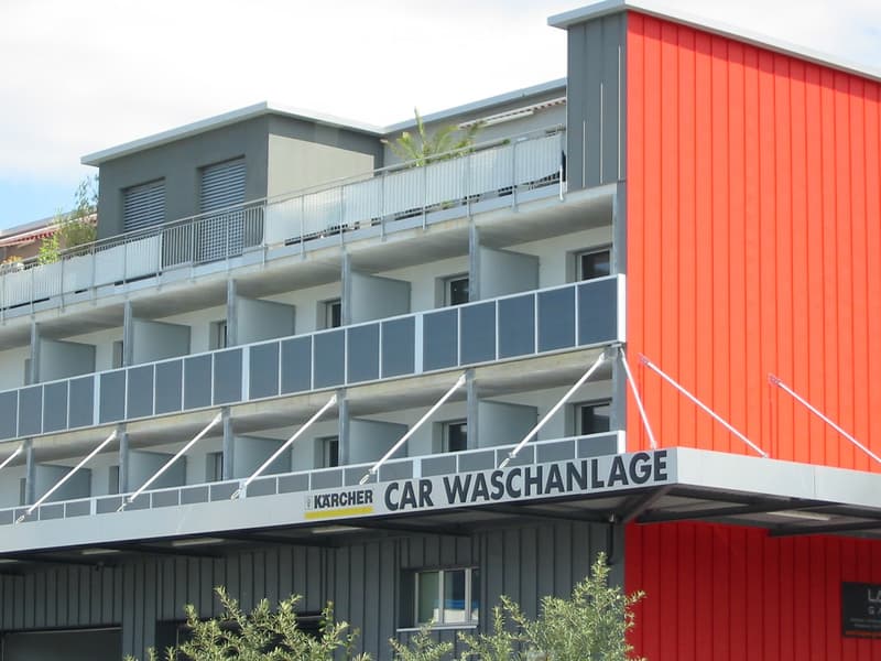 Modern multi-story building with balconies, red and grey exterior, and a sign reading "CAR WASCHANLAGE" (car wash).