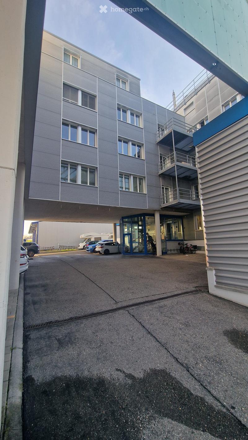 Modern multi-story building with grey facade, large windows, and a covered entrance area. Several cars are parked in front, and a few bicycles are near the entrance.