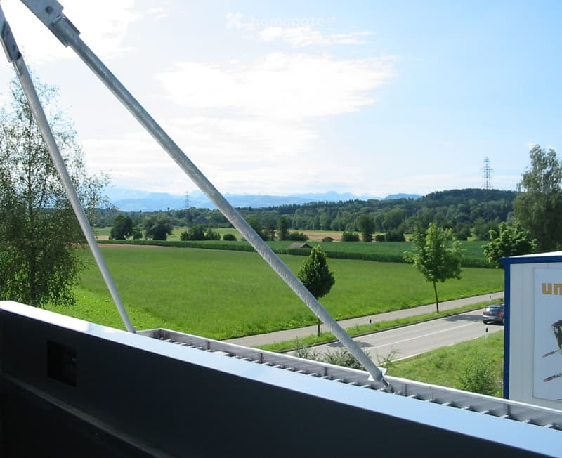 Balcony overlooking a green field with trees, a road, and distant mountains under a partly cloudy sky.