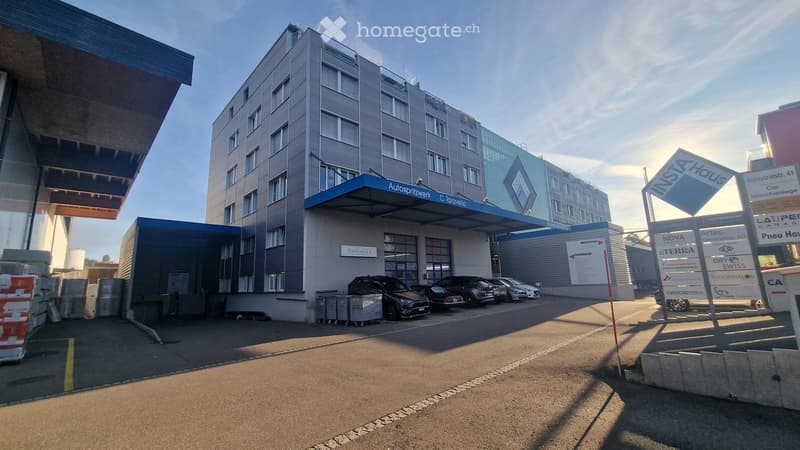 Modern multi-story building with a blue awning and signage for 'Autosportwerk' and 'C. Taravella'. Several cars are parked in front, and there are various commercial signs visible.