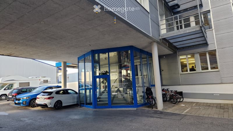 Modern building entrance with glass doors, several parked cars, and bicycles near the entrance.