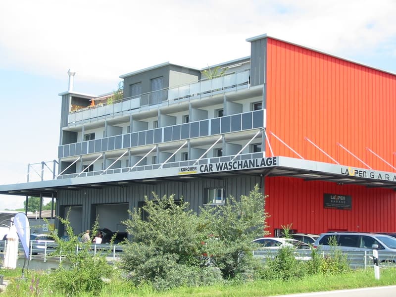 Modern multi-story building with red and grey exterior, featuring balconies, signage for "CAR WASCHANLAGE" and "LAOPEN GARAGE", and several parked cars in front.