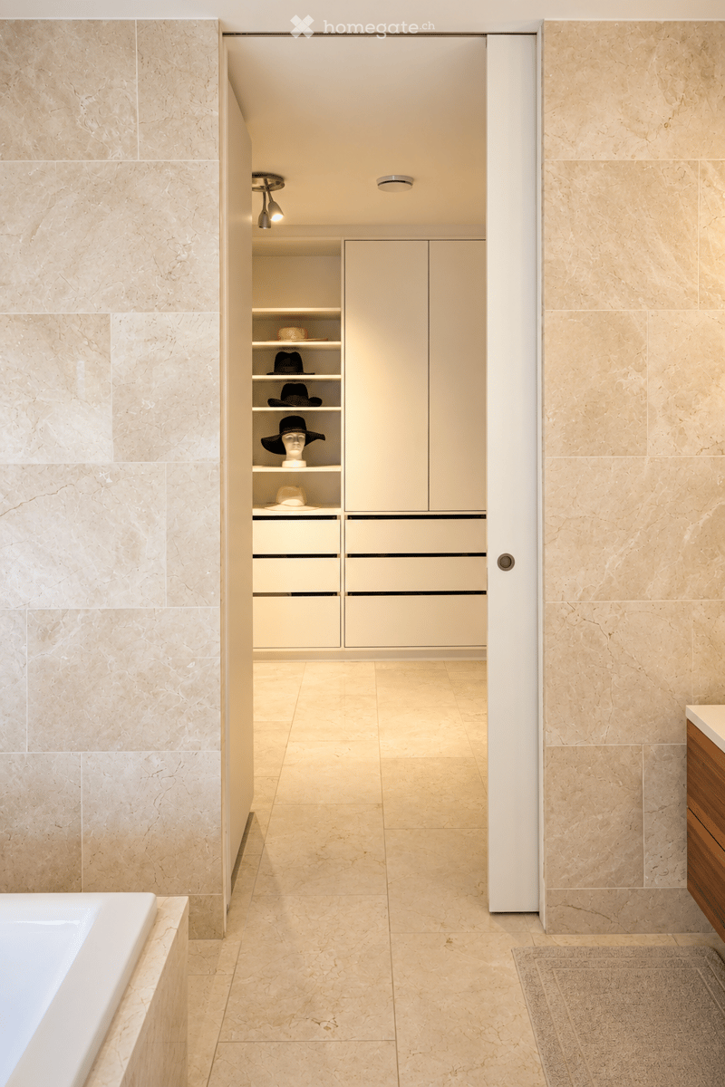 Modern bathroom with beige marble tiles, a white bathtub, built-in storage cabinet, and decorative hats on shelves.