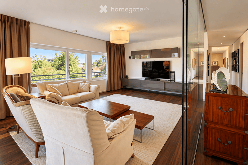 Modern living room with large windows offering a scenic view, featuring a cream-colored sofa, wooden coffee table, armchair, and a television mounted on the wall.
