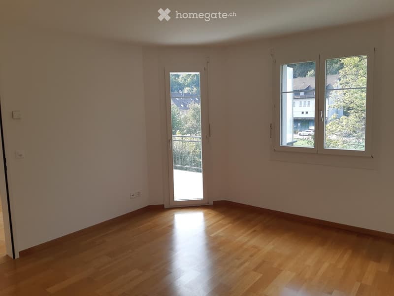 Empty room with wooden flooring, white walls, and large windows with a view of the outside. A sliding door leads to a balcony.