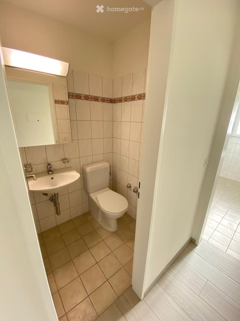 Small bathroom with white tiles, toilet, sink, and mirror. Beige tiles on the floor.