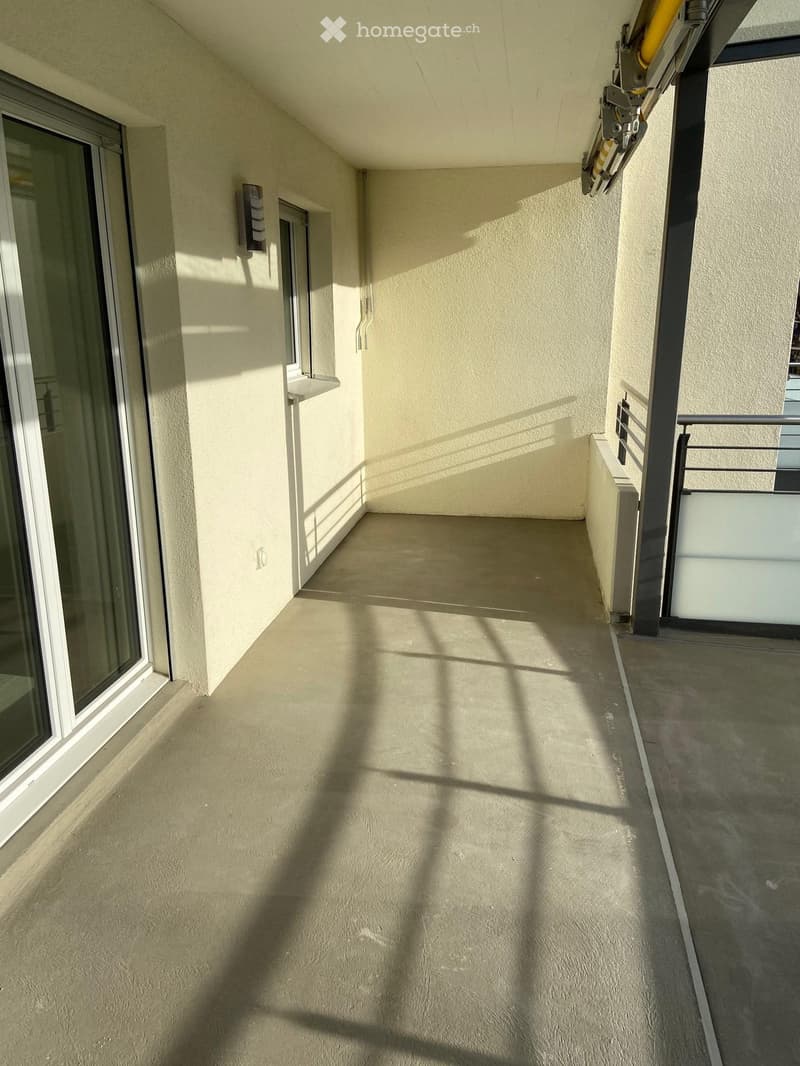 An empty balcony with a concrete floor, white walls, and a glass door. There is a wall-mounted light and a window on the left side.