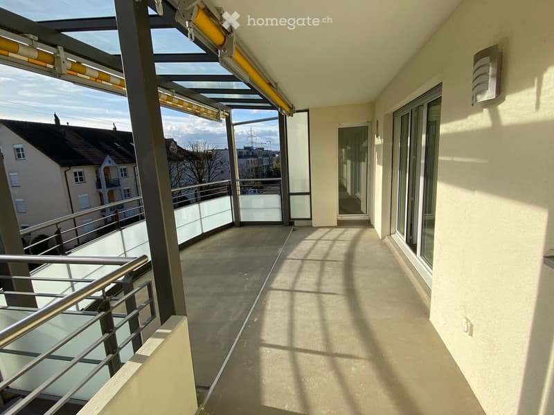 Empty balcony with glass railings, metal support beams, and a glass roof. The floor is made of concrete. The wall is painted in a light color. There is a wall-mounted light on the right side.