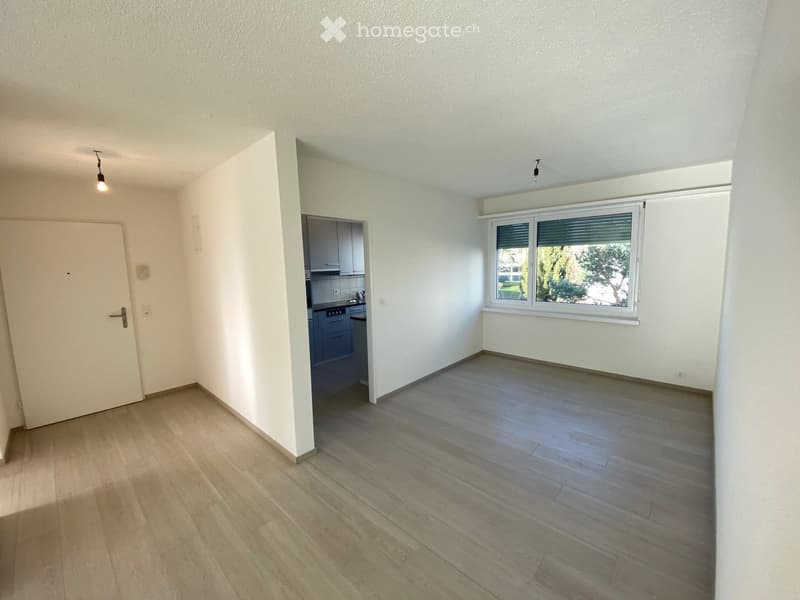 Empty room with white walls, wooden floor, and a door leading to a kitchen. A window with blinds is on the right side.