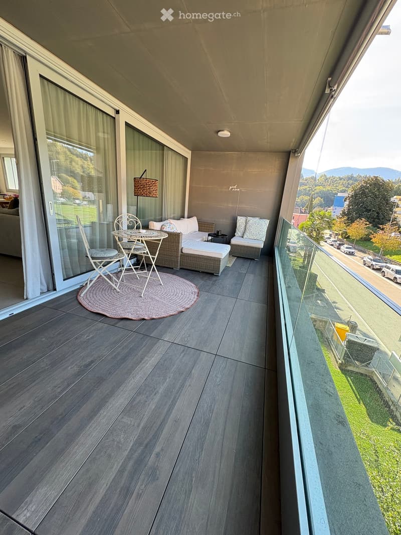Balcony with wooden flooring, sofa, small table, chairs, and a view of the street and surrounding landscape.