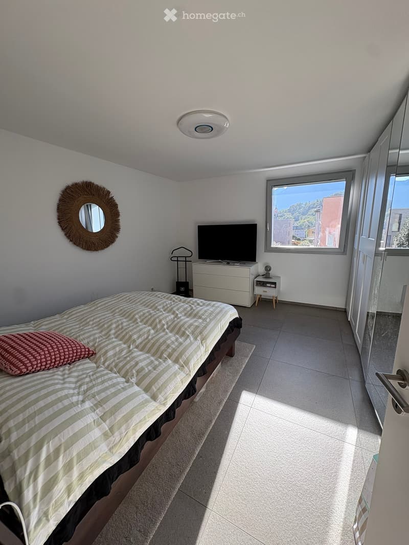 Modern bedroom with a neatly made bed, a large window offering a scenic view, a television on a white stand, a round mirror on the wall, and a small nightstand with a lamp.