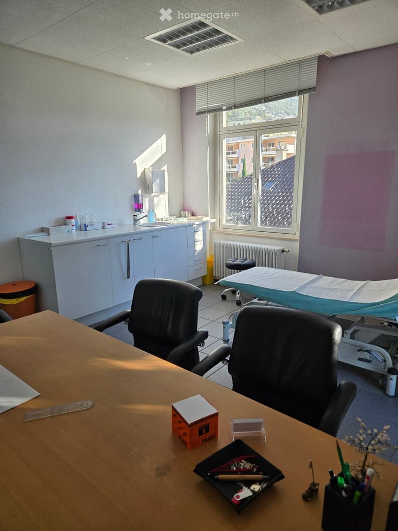 A well-lit room with a large window, a wooden table with chairs, a sink area with cabinets, an exam table, and a radiator. The room also includes a trash bin and a stool.