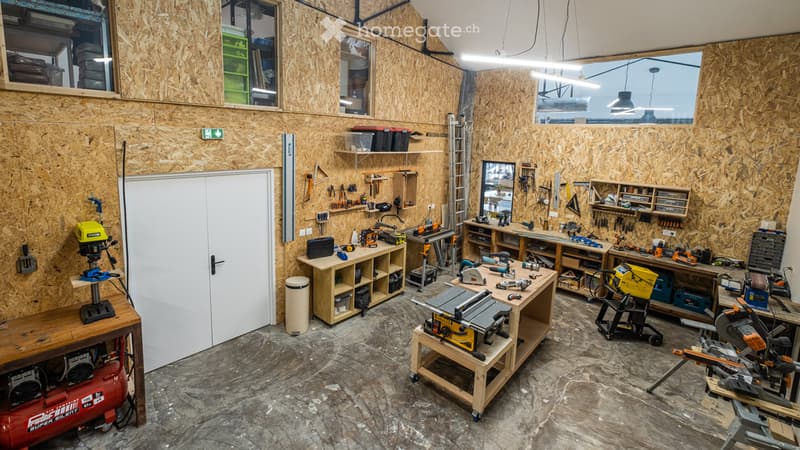 Well-organized workshop with wooden walls, concrete floor, and large windows. Contains workbenches, tools, storage shelves, and equipment like a table saw and air compressor.