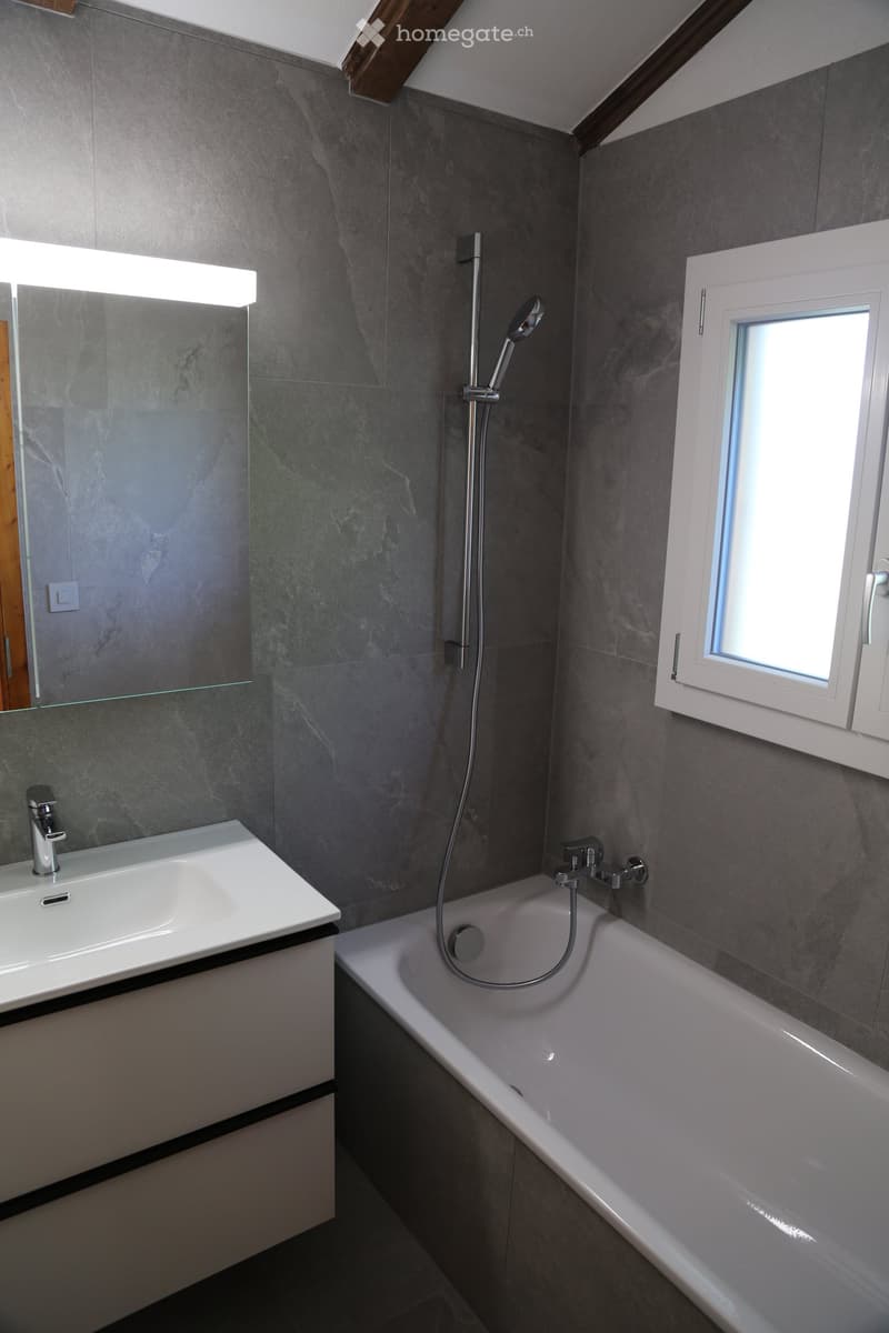 Modern bathroom with a bathtub, sink, and showerhead. Grey marble tiles on walls, a large mirror with built-in lighting, and a small window letting in natural light.