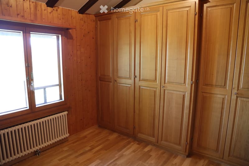 Empty room with wooden walls and flooring, large window, built-in wooden cabinets, and a radiator.